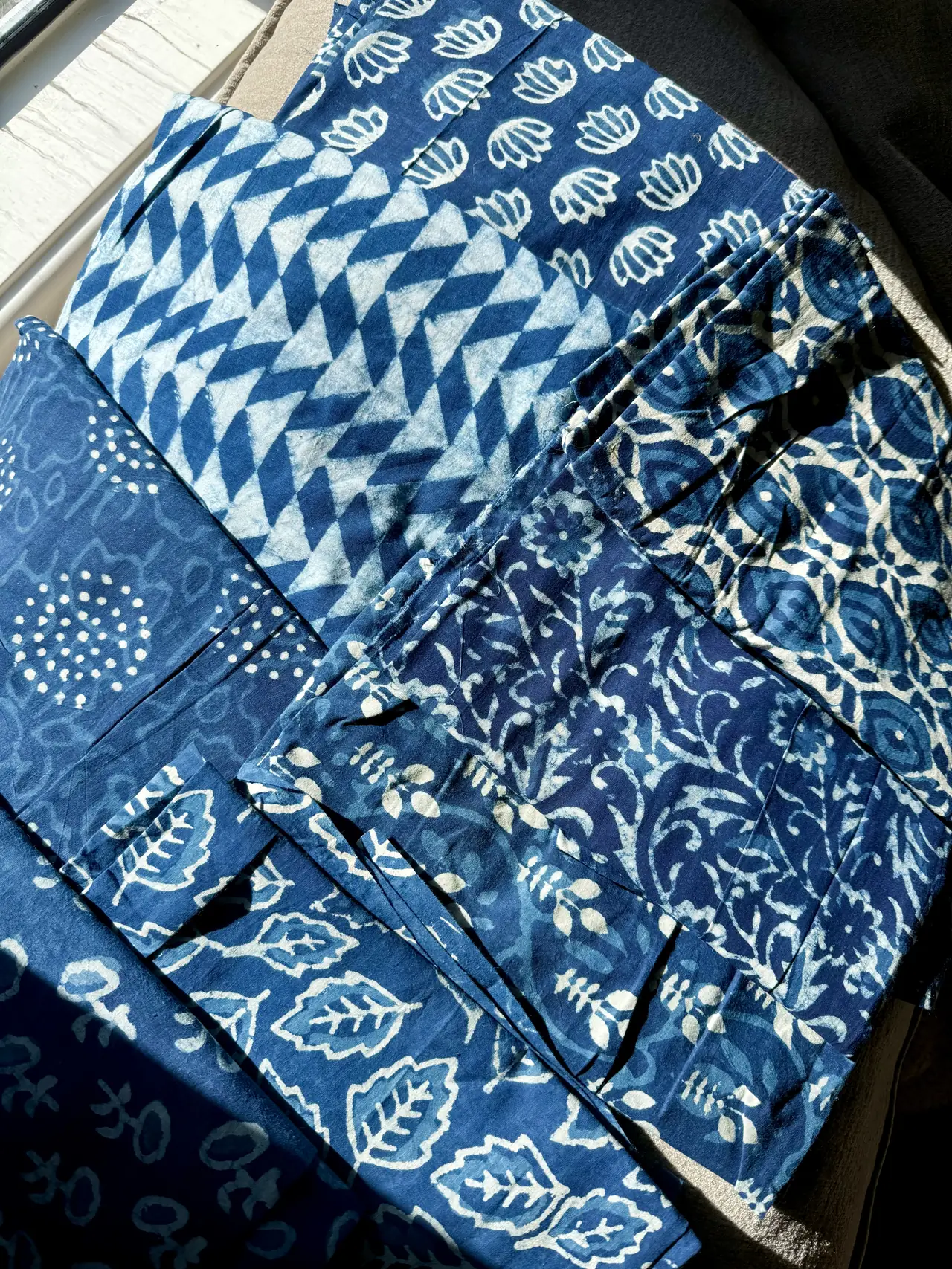 Blue and cream fabrics with floral, leaf, dot, and geometric patterns, spread together beside a window