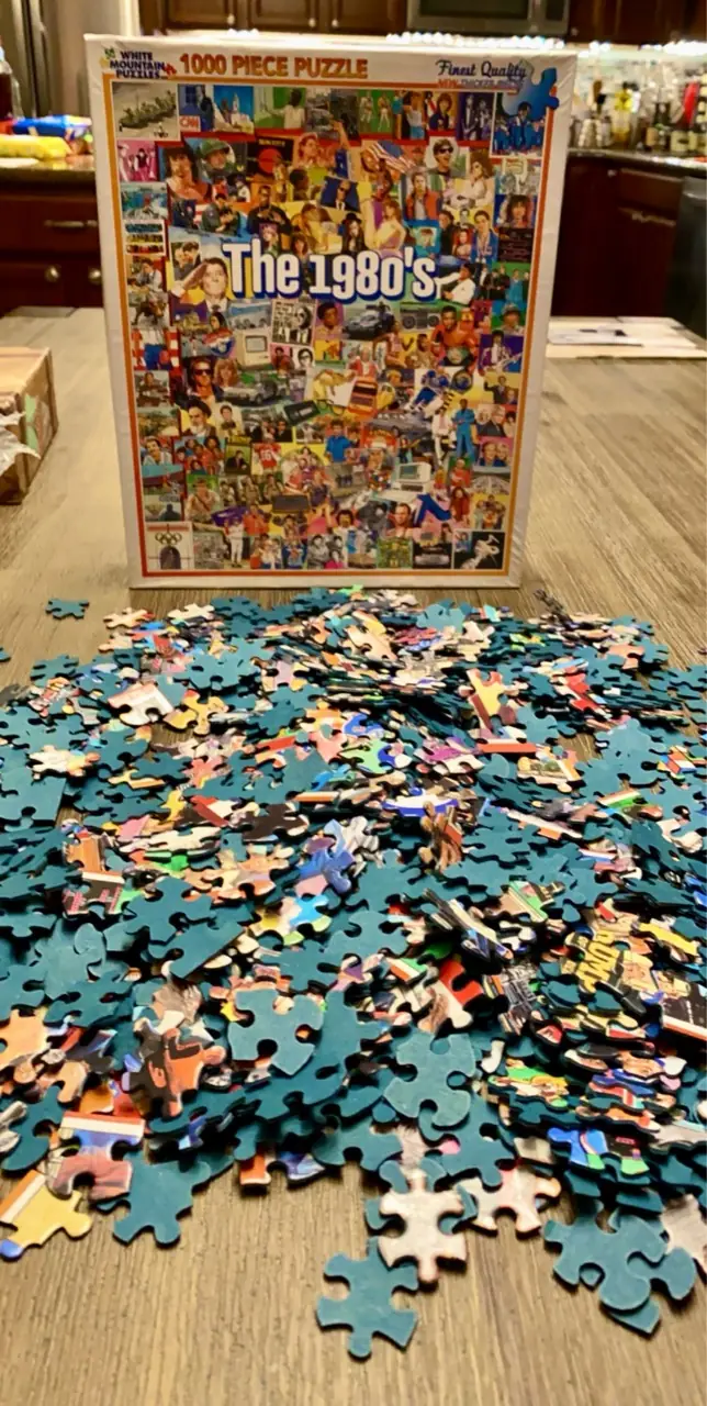 A White Mountain Puzzles The 1980s box behind loose puzzle pieces spread across a table