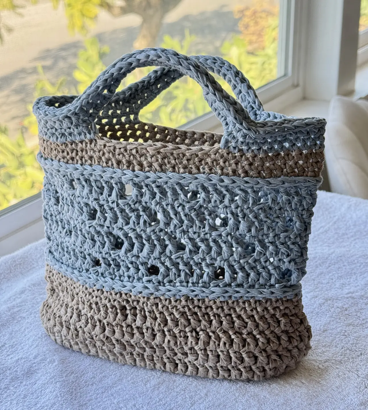 A crocheted market bag in blue and taupe yarn beside a window