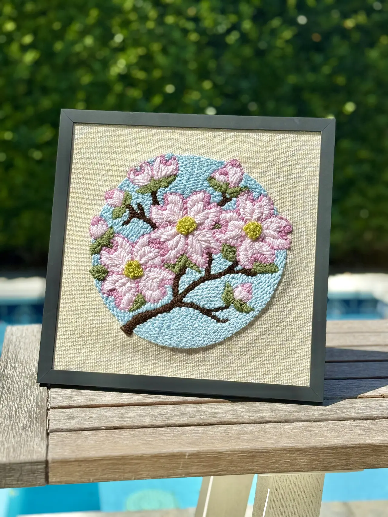 A framed punch needle picture of pink blossoms against a pale blue sky