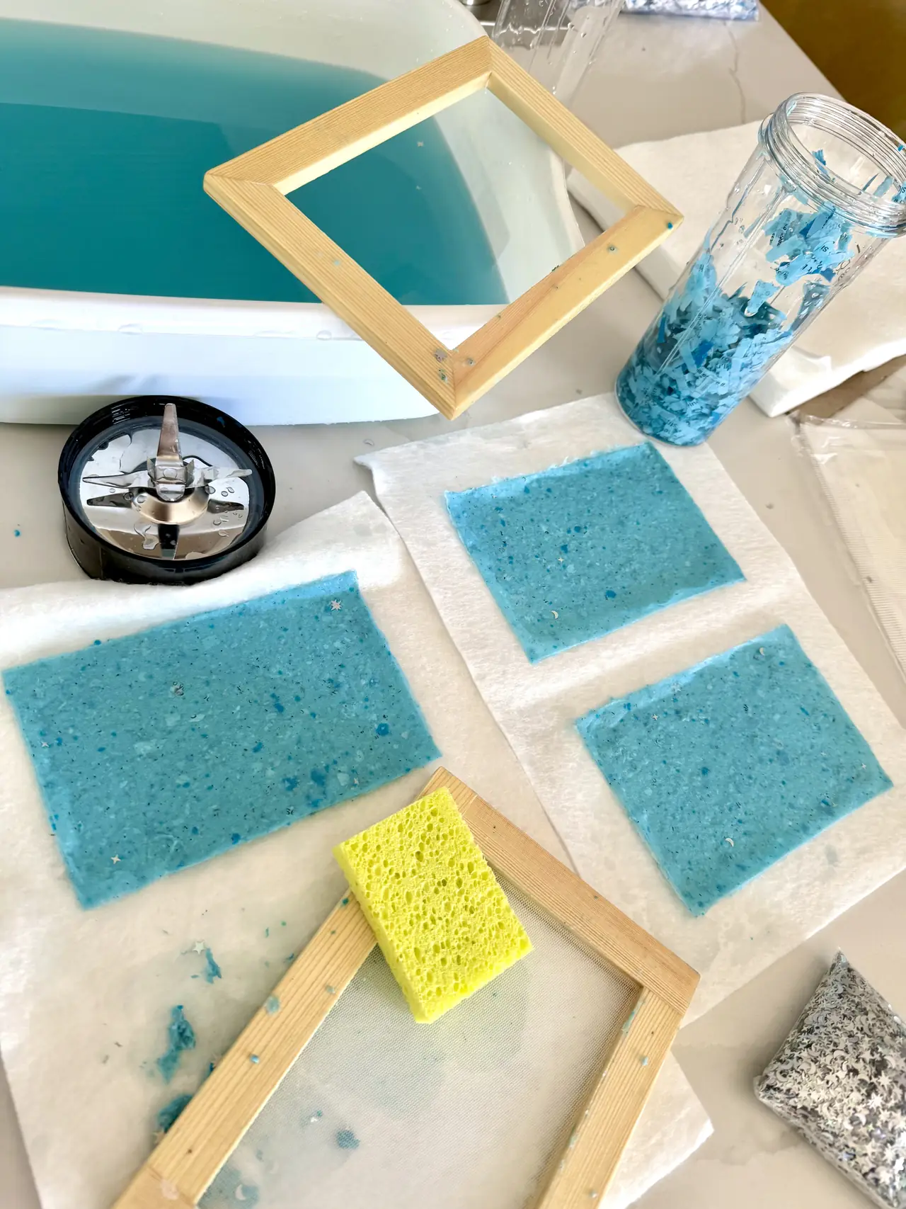 Sheets of handmade blue paper drying beside a wooden mould and a jar of pulp